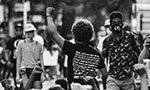Foto em preto e branco. No centro uma pessoa com cabelo afro, camiseta preta e punho erguido