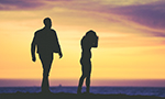 Fotografia da silhueta de um casal ao por do sol, a mulher está com a cabeça baixa, escondendo o rosto com as braços e o homem está andando em sua direção.