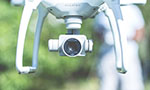 Um drone branco com uma câmera no centro e o texto "PHANTOM" na frente. Atrás, fundo verde desfocado e uma pessoa em pé usando roupas brancas.