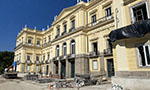 Fachada do Fachada do Museu Nacional/UFRJ, no Rio de Janeiro, com materiais de construção na frente dispostos no chão. Há também pessoas trabalhando na obra e um caminhão. É um prédio de três andares em tom amarelo pastel. Elementos da arquitetura imperial: arco, alto relevo, janelas retangulares, janelas com arcos, topo triangular.