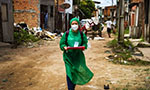 Foto em forma retangular horizontal. Mulher usando equipamento de proteção (máscara branca, touca verde, uma espécie de avental que cobre os braços, o corpo e as pernas em tom verde). Ela está andando em direção à câmera e está no centro da imagem. Ela usa mochila preta, um crachá branco pendurado no pescoço e segura uma pasta e uma caneta. No fundo, uma rua de terra batida com casas simples, muita sujeira, uma carroça, uma pessoa na frente de um portão e outra do lado da carroça.
