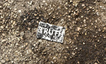 Foto retangular horizontal. Papel retangular na horizontal com a palavra TRUTH (verdade, em inglês) muito desgastado no chão. O papel é preto ou cinza escuro e as letras brancas. No chão pedregulhos e terra.