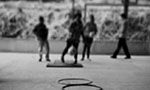 Foto em preto e branco borrada, formato retangular e na vertical. Uma quadra de escola, quatro crianças em pé no fundo. No chão, bambolês dispostos um na frente do outro fazendo uma linha.