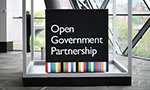 Foto de um quadro preto com o texto "Open Government Partnership". Uma faixa listrada e colorida na parte de baixo. O quadr está dentro de um aquário vazio e retangular. No fundo, uma sala ampla e clara com paredes envidraçadas.