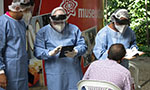 Foto externa. Três pessoas em pé com avental de corpo inteiro, luvas, máscara, protetor facial de acrílico e touca. Uma usa um tablet, outra uma prancheta com folhas. Na frente, um homem sentado. Atrás, um poster do Museu da Vida (Fiocruz).