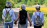 Foto: três mulheres de costas, andando em uma trilha de mãos dadas. No fundo, um espaço verde com árvores e grama.