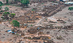 Foto aérea tirada em Mariana, Minas Gerais (2015) após o rompimento de uma barragem. A foto mostra uma região plana coberta por lama, casas destruídas, árvores isoladas e um helicóptero sobrevoando.
