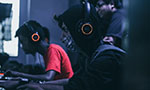 Foto: Dois homens jovens com fones de ouvido e máscara. Também é possível ver a sombra de uma terceira pessoa. Eles estão concentrados em seus computadores. A sala parece estar um pouco escura. Um outro rapaz está em pé atrás dos jovens olhando para uma das telas.