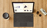 Foto ou montagem de um notebook sobre mesa de madeira. Na tela, uma página do Word e um site de câmeras. No lado esquerdo do computador, um copo descartável de café. No lado direito, uma câmera e um cilindro preto com a tampa aberta.