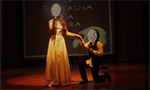 Foto de peça de teatro encenada por duas crianças. Uma menina usando um vestido que vai até os pés segurando microfone. Um menino com roupa social segura um microfone. Ele está com um joelho no chão e segurando a mão da menina.