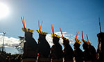 Foto da Marcha das Mulheres Indígenas, Brasília - DF (2019). Oito mulheres indígenas de costas. É possível ver apenas a silhueta e seus cocares. No fundo, céu azul com poucas nuvens, o sol, algumas árvores e um grupo de pessoas.