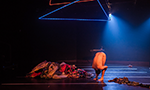 Foto do espetáculo “Strip Tempo – Stripteases contemporâneos”. Palco escuro com um foco de luz azul e duas luminárias geométricas de led no teto. Uma pessoa está performando no centro do palco. Ela está em pé, com a cabeça para baixo, totalmente curvada e abraçando as pernas. Do lado esquerdo, uma pilha de roupas bagunçadas.