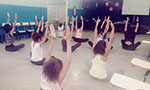 Foto de uma sala de aula. As cadeiras foram colocadas próximas das paredes. Um grupo de alunos está sentado no chão e de costas para a câmera. Eles estão de pernas cruzadas e com os braços para cima.