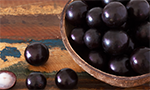 Foto de um recipiente cheio de jabuticaba. Do lado, três jabuticabas inteiras e três metades com a polpa para cima. Mesa de madeira.