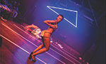 Foto de uma performance de striptease. Uma mulher está se apresentando. Há uma plateia no canto esquerdo. Ela está em um espaço aberto que serve como palco, há alguns objetos amontoados no fundo. Ela está com as mãos sobre o peito e olha para trás.