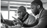 Dois homens negros sentados diante de um computador. Ambos, o mais velho e o mais novo, estão apontando para o computador. A expressão facial dos dois é de seriedade e concentração.