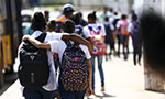 Foto de duas jovens usando mochila. Elas estão de costas, andando na rua e com o braço sobre o ombro uma da outra. Mais a frente, outros jovens estão caminhando.