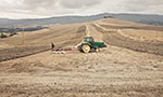 Foto de um trator no meio de um campo meio amarelado. No fundo, relevo montanhoso.