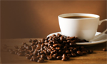 Xícara cheia de café repousando sobre um pires branco, posicionado sobre uma mesa de madeira. Espalhados ao redor da borda do pires e sobre a superfície da mesa, encontram-se grãos de café torrados.