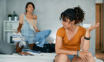 Duas mulheres estão discutindo. A que está mais à frente, sentada em uma cama, está olhando de perfil para a outra, que está em pé, mais ao fundo. Ela segura algumas roupas, com expressão de raiva, enquanto encara a primeira mulher.