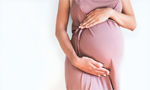 Fotografia de uma mulher grávida que está em pé, usando um vestido rosa, com as mãos sobre sua barriga.