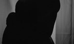 Fotografia de uma mulher grávida em pé em frente a uma cortina. Ela está de perfil e com as mãos na barriga. A imagem escura não permite que seu rosto seja visto com nitidez, apenas sua silhueta.