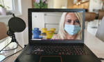 Fotografia de um notebook ligado, exibindo uma mulher loira com máscara na tela. O dispositivo repousa sobre uma mesa branca espelhada, ao lado de um microfone de mesa. Ao fundo, há uma sala ampla com alguns móveis, que está desfocada.