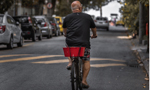 Idoso desfrutando de um passeio de bicicleta pelo meio de uma rua arborizada, ladeada por árvores dos dois lados e com alguns carros. Na fotografia, ele direciona seu olhar para uma das árvores, no lado direito.