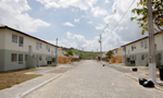 Fotografia de um condomínio do programa de habitações populares "Minha Casa, Minha Vida". Nela é possível ver uma rua ladeada por edifícios de um andar, que alternam-se entre edifícios cinza e branco e edifícios laranja e branco. Ao fundo da imagem, há um morro com algumas vegetações.