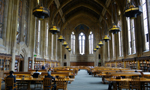 Interior de uma biblioteca da Universidade de Washington, em Seattle, mais conhecida como Suzzallo Library. Na fotografia, há uma grande sala de leitura com mesas e cadeiras de madeira, onde algumas pessoas estudam. A sala é ladeada por prateleiras de livros.