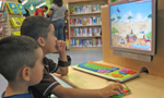 Dois meninos em uma biblioteca jogando em um computador. Ao fundo, há prateleiras com livros, ao lado de duas mulheres adultas, que estão em pé.
