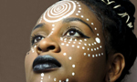Mulher negra com brincos de argola, cabelo preso em coque, usando colar preto e batom preto. Seu corpo está coberto por pintura corporal branca. Ela olha para o horizonte.