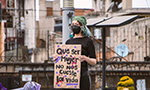 Mulher de cabelos curtos com mechas azuis, usando máscara e roupa preta, segurando uma placa durante um manifesto contra a violência às mulheres. Na placa está escrito: "que ser mujer no nos cueste la vida" [que ser mulher não nos custe a vida, em português].