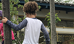 Menina de costas, usando tênis preto, calça azul marinho e blusa de manga longa branca e cinza, se apoia em uma árvore com a mão esquerda para subir um degrau. Mais adiante, uma menina vestindo agasalho rosa e touca de crochê lilás caminha em direção a uma escola visível ao fundo da fotografia.