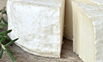 Queijo inteiro sobre uma mesa de madeira com um ramo de alecrim ao lado, apresentando uma fatia grossa cortada que sobressai da peça.