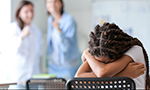 Jovem negra com tranças apoiando a cabeça nos braços sobre a parte de trás de uma cadeira na sala de aula, enquanto duas garotas, desfocadas ao fundo, parecem ridicularizá-la.