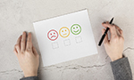 Duas mãos sobre uma superfície cinza com rachaduras, segurando uma caneta para preencher um papel que está logo à sua frente. No papel, há três caixas para marcação, acompanhadas por emojis representando diferentes expressões faciais: insatisfação, neutralidade e satisfação, da esquerda para a direita.