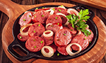 Panela rasa de ferro preta com linguiças fatiadas, anéis de cebola e cheiro verde, sobre uma tábua de madeira. Ao lado, um recipiente com óleo e outra tábua com pimentas coloridas, tudo disposto sobre uma mesa de madeira escura e rústica.