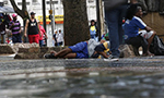 Fotografia de pessoas caminhando perto de uma pessoa deitada no chão e outra sentada perto de uma árvore em uma área urbana movimentada em um dia chuvoso.
