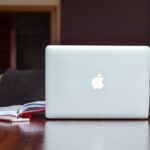Fotografia de um caderno aberto com um celular em sua frente e um computador ao lado, sobre uma mesa