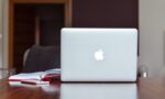 Fotografia de um caderno aberto com um celular em sua frente e um computador ao lado, sobre uma mesa