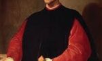 Retrato de Nicolau Maquiavel. Ele está de pé em um fundo com uma parede marrom. É branco e usa roupas pretas e vermelhas.