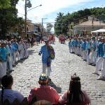 Fotografia da Congada, nas ruas de Ilha Bela. Há homens enfileirados na esquerda e na direita e um homem no centro, todos usando camisas azuis e calças brancas. Na frente dos homens, há o público