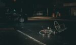 Fotografia de uma rua escura com um carro batido e uma bicicleta caída no chão
