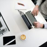 Fotografia de uma mesa branca com um notebook, ao qual um homem branco e de camisa de botões cinza está digitando, um bloco de anotações em branco com duas canetas, uma xícara com café, um tablet, uma calculadora, um controle remoto e um aparelho de som