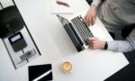 Fotografia de uma mesa branca com um notebook, ao qual um homem branco e de camisa de botões cinza está digitando, um bloco de anotações em branco com duas canetas, uma xícara com café, um tablet, uma calculadora, um controle remoto e um aparelho de som