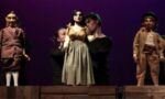 Reprodução do 5º Canto de Auto da Catingueira. Três marionetes, duas representando mulheres e um representando um homem, com seus marionetistas atrás. As marionetes estão de pé em um palco de madeira com um fundo escuro