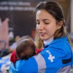 Fotografia de uma pessoa vestindo um uniforme azul do SUS segurando um bebê nos braços.