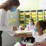 Fotografia de uma professora medindo a temperatura de uma aluna em uma sala de aula.